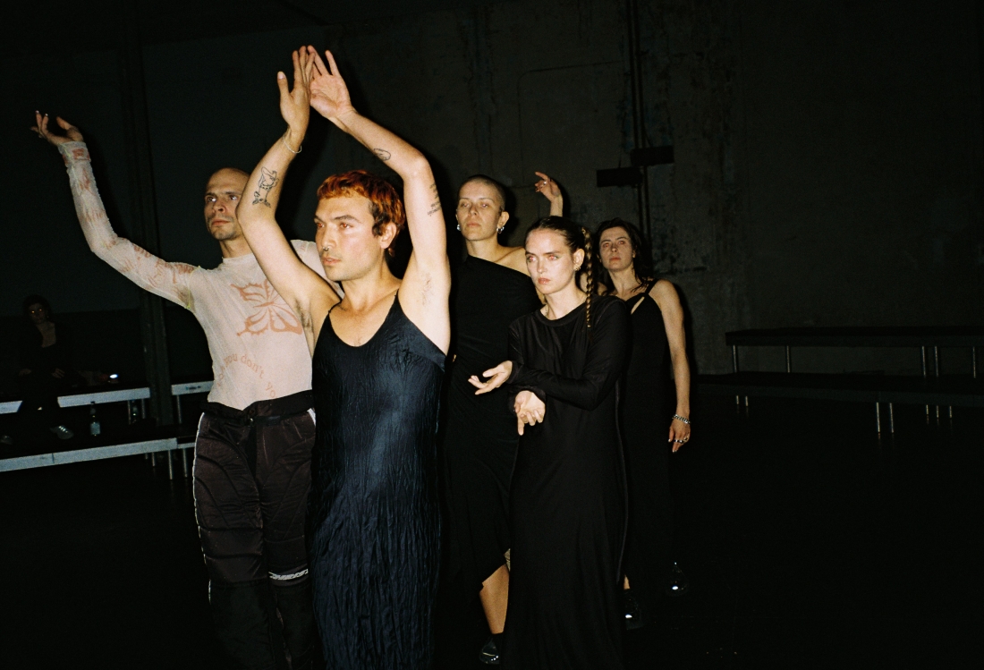 Color scene photograph. A performer wearing a short black dress raises his arms.