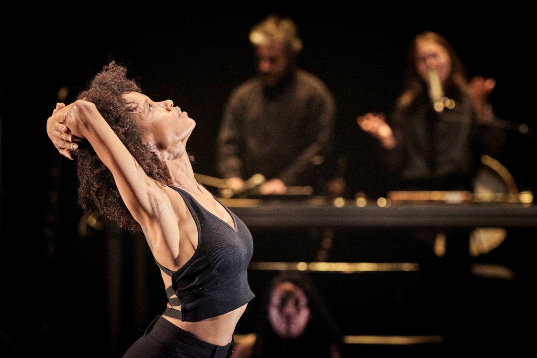   An performer stretches her arms behind her head in the foreground, whilst two other performers, blurred in the background, stand by a table.