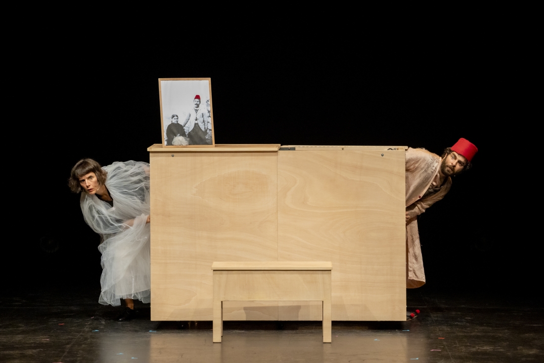 Two people peer out from opposite sides of a large wooden piece of furniture on stage. On top of the furniture is an old photograph with red details.
