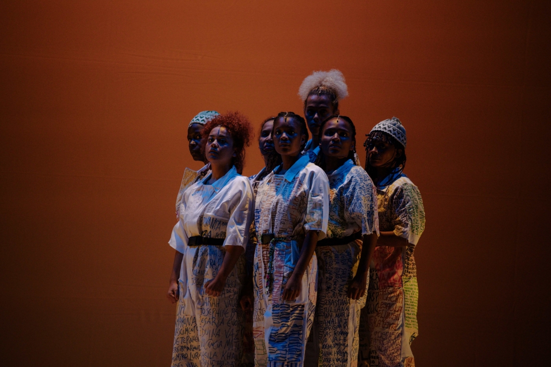 Color stage photo. At the center of the action, seven black female performers have the same type of clothing: a white suit, from top to bottom and with several names inscribed. With their fists clenched, they form a group.