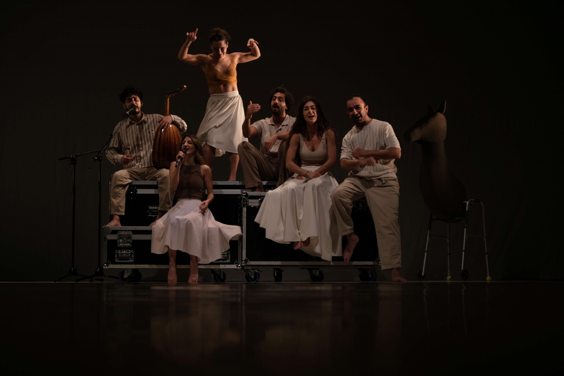 Six barefoot performers sing and play music whilst sitting on stage transport crates, whilst a performer dances standing in the centre.