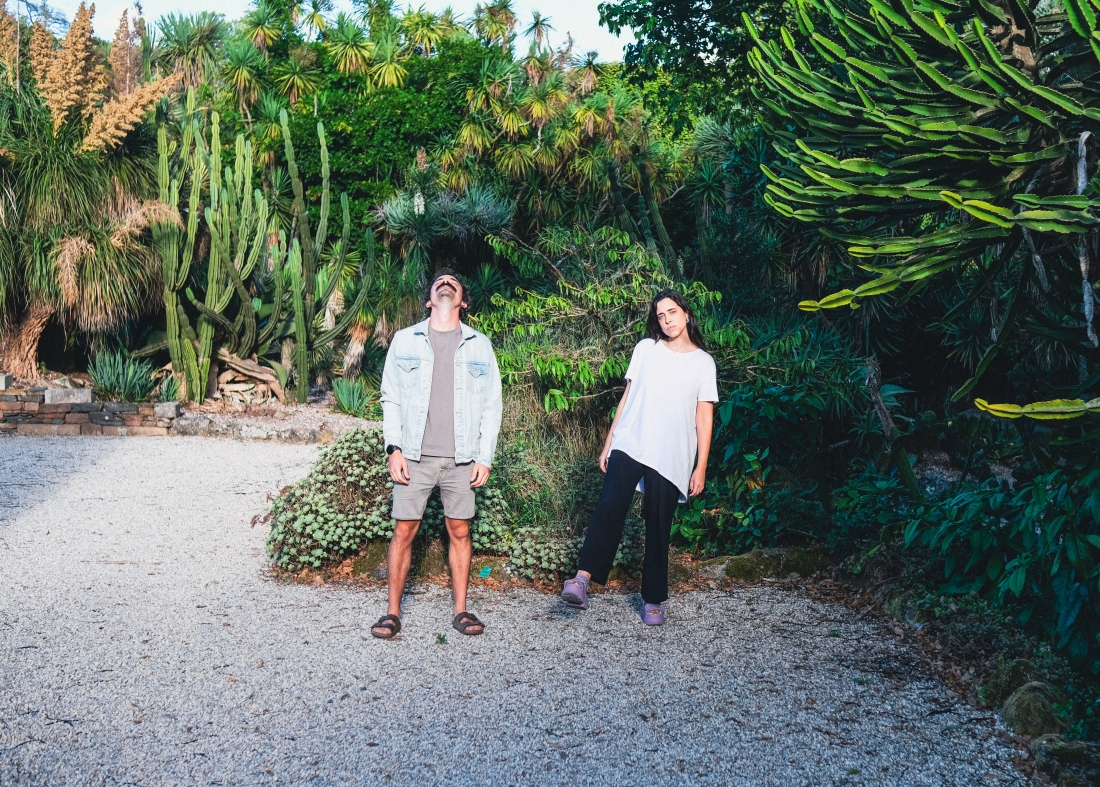 Sofia and José are standing side by side in front of a cluster of tall cacti, looking at the camera with neutral expressions.