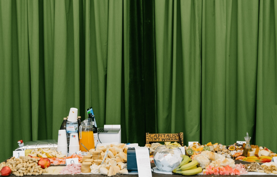 Color photo. On a table, lots of food, juice, a bottle of water, a computer and a piece of cardboard that reads: “Capitalism isn't working.”