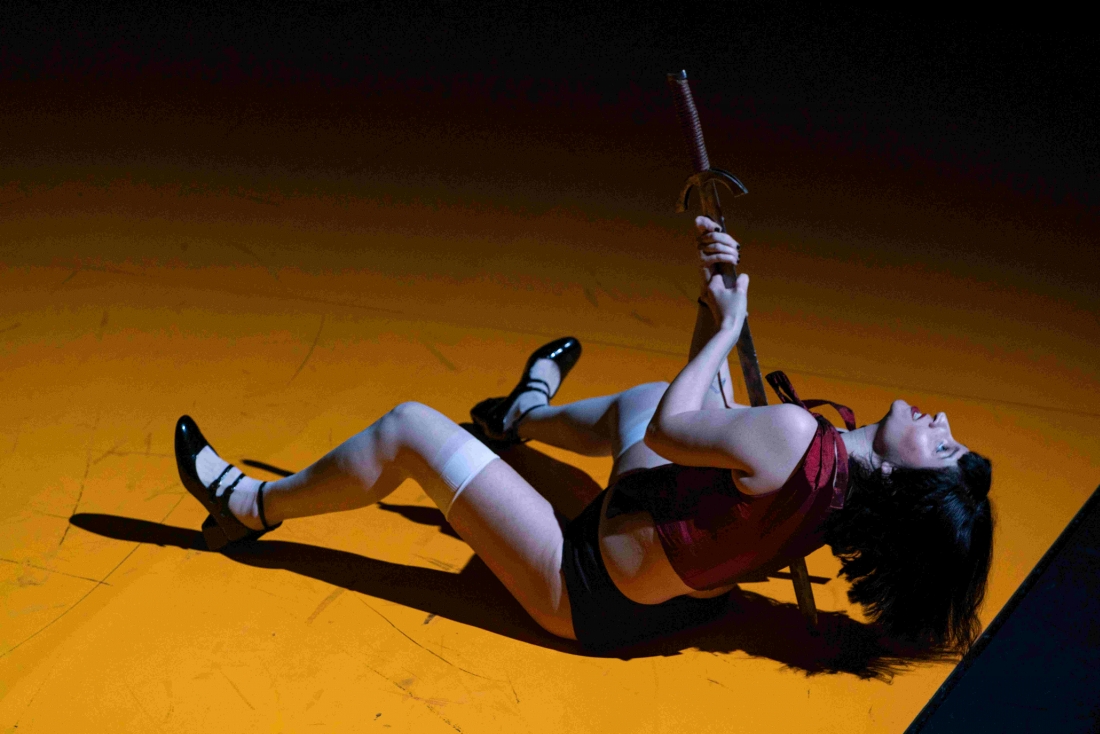 A woman is lying on the floor of a stage, holding a sword upright by her side. She is wearing a red top, black shorts, white knee-length socks and black shoes, standing out against the illuminated yellow floor.
