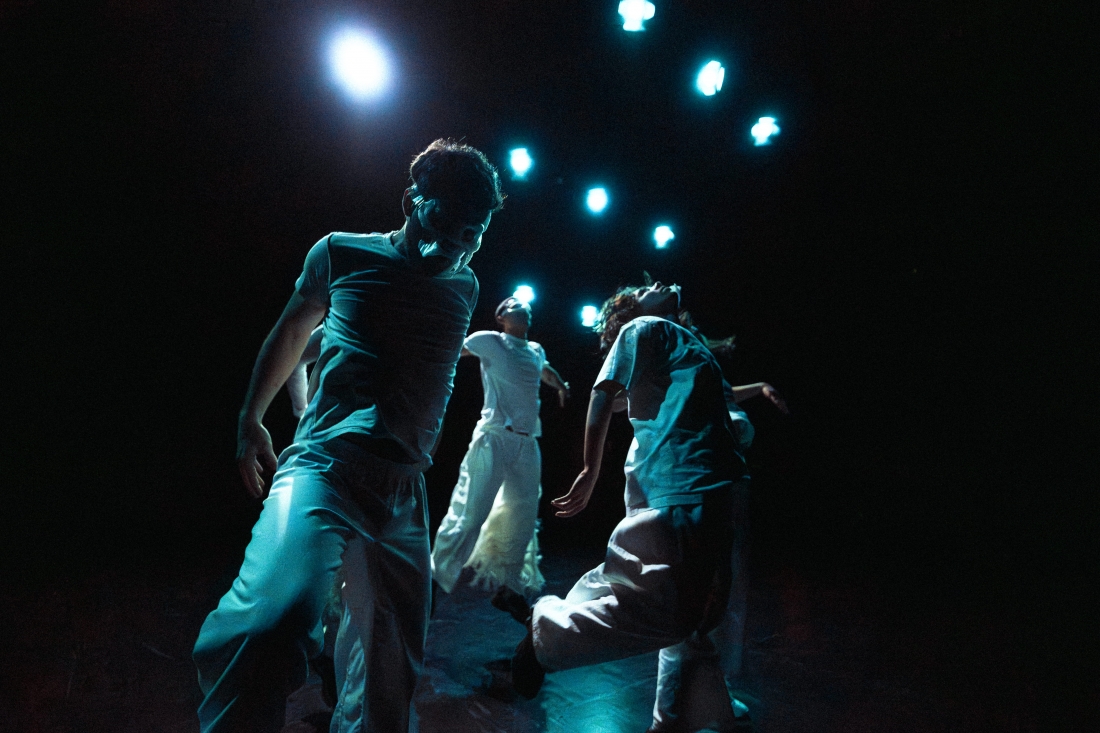 Color photograph of a man's face in profile. Part of his face is covered by a skull-shaped helmet, and he is playing a bone-like wind instrument.