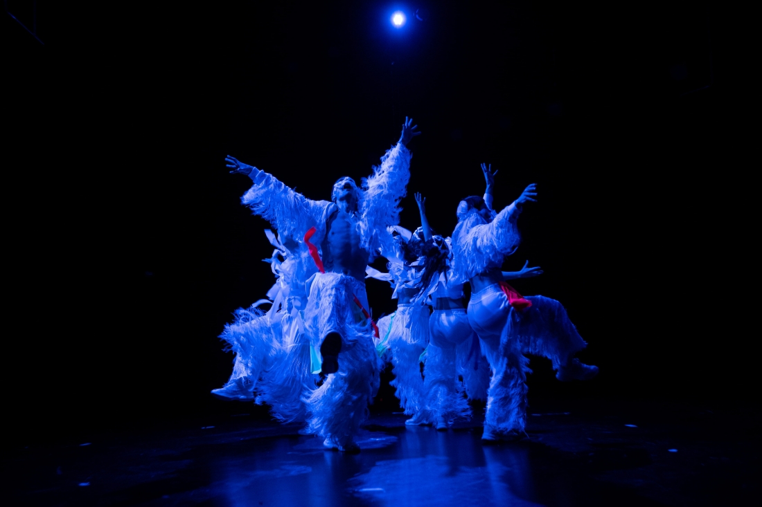Fotografia a cores do rosto de um homem, de perfil. Parte do seu rosto está coberta por um capacete em forma de esqueleto e ele toca um instrumento de sopro semelhante a um osso.