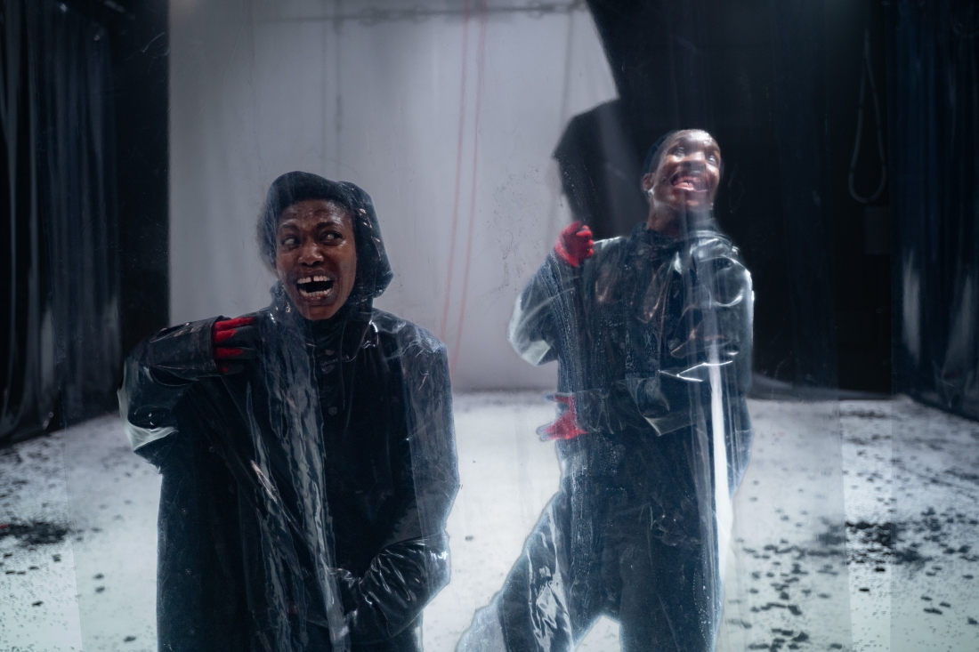 Fotografia de cena a cores. Dois intérpretes com expressões faciais horríficas, atrás de uma tela de plástico. Vestem parcas pretas e luvas vermelhas.