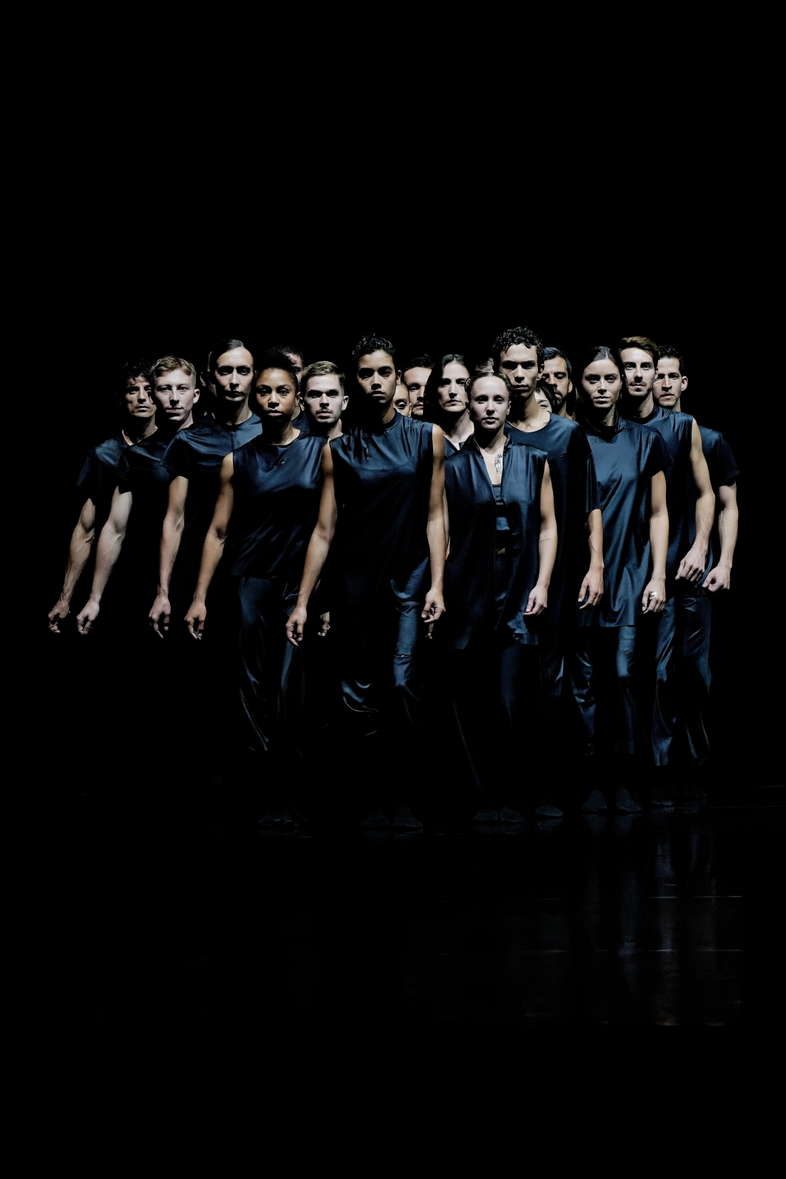 Fotografia de cena a cores de um grupo de bailarinos e bailarinas vestidos de preto, com figurinos muito semelhantes, movendo-se em sintonia na penumbra, como um único organismo.
