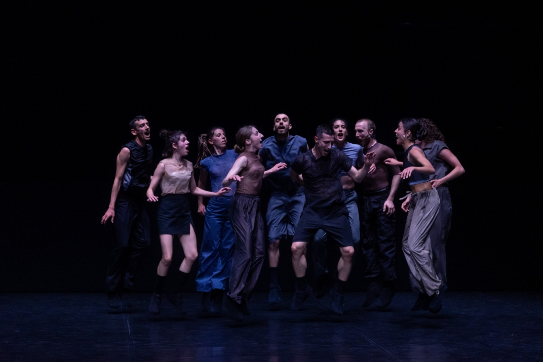 Color stage photograph of a group of ten performers jumping and vocalizing together, dressed in shades of blue, gray, white, and black.