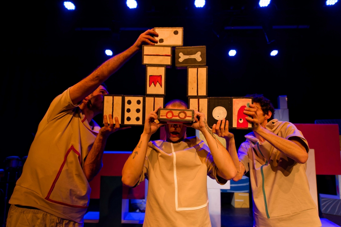 Two performers on stage stack small cardboard blocks with illustrations around a third performer in the centre, who holds a block in front of his eyes as if they were glasses.
