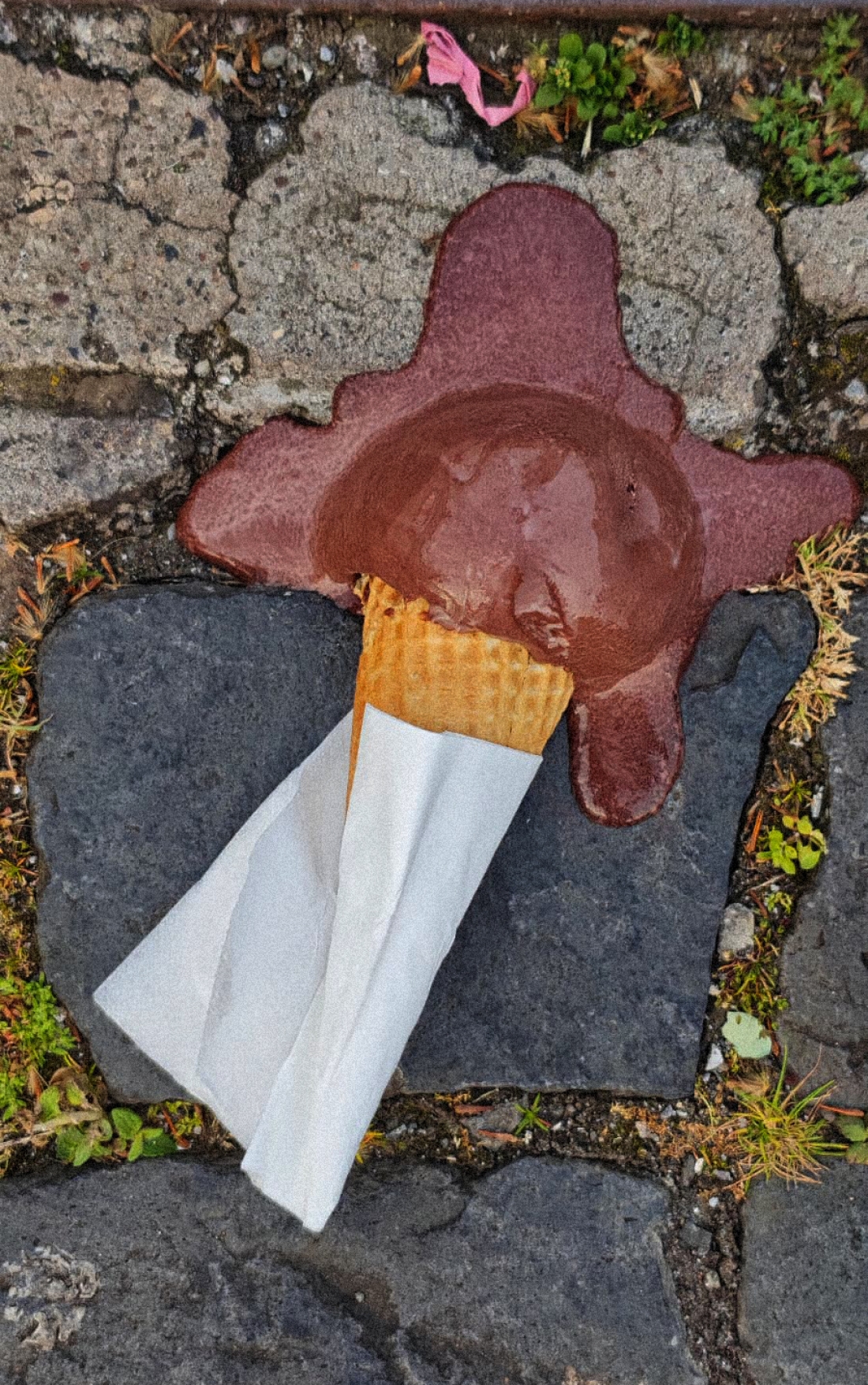 Um gelado de chocolate num cone, envolto num guardanapo, derrete numa calçada da rua.