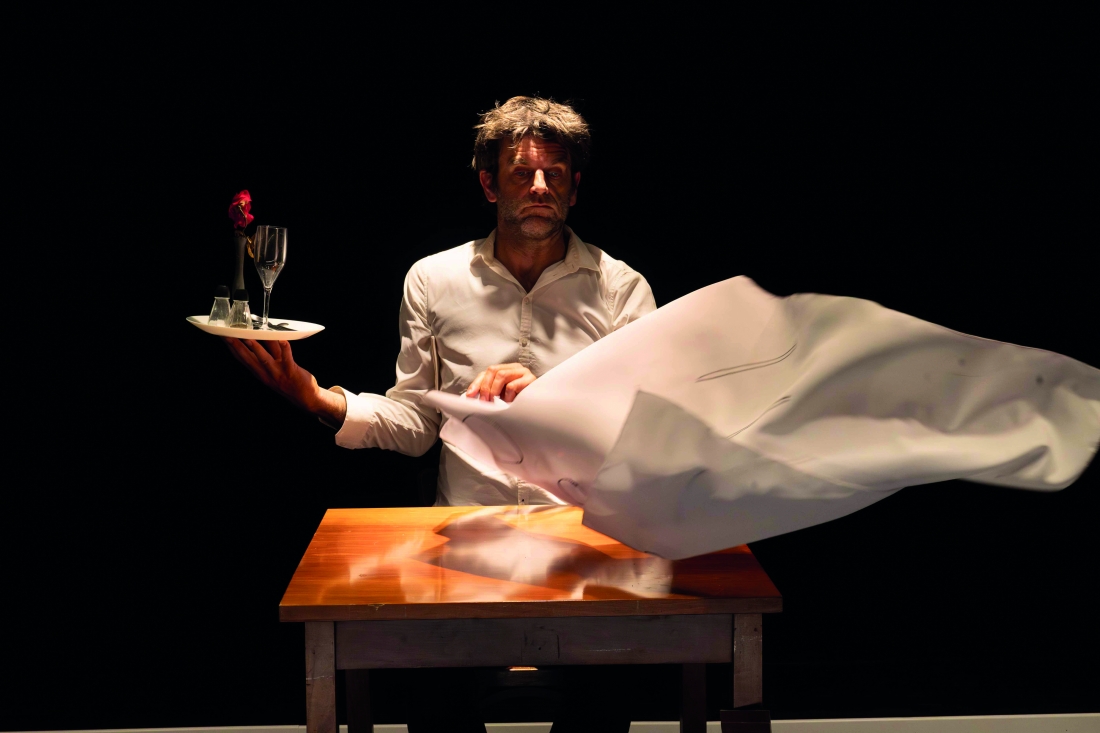 A man is sitting at a table, holding a tray with glasses and a flower, whilst a white cloth flutters in front of him.
