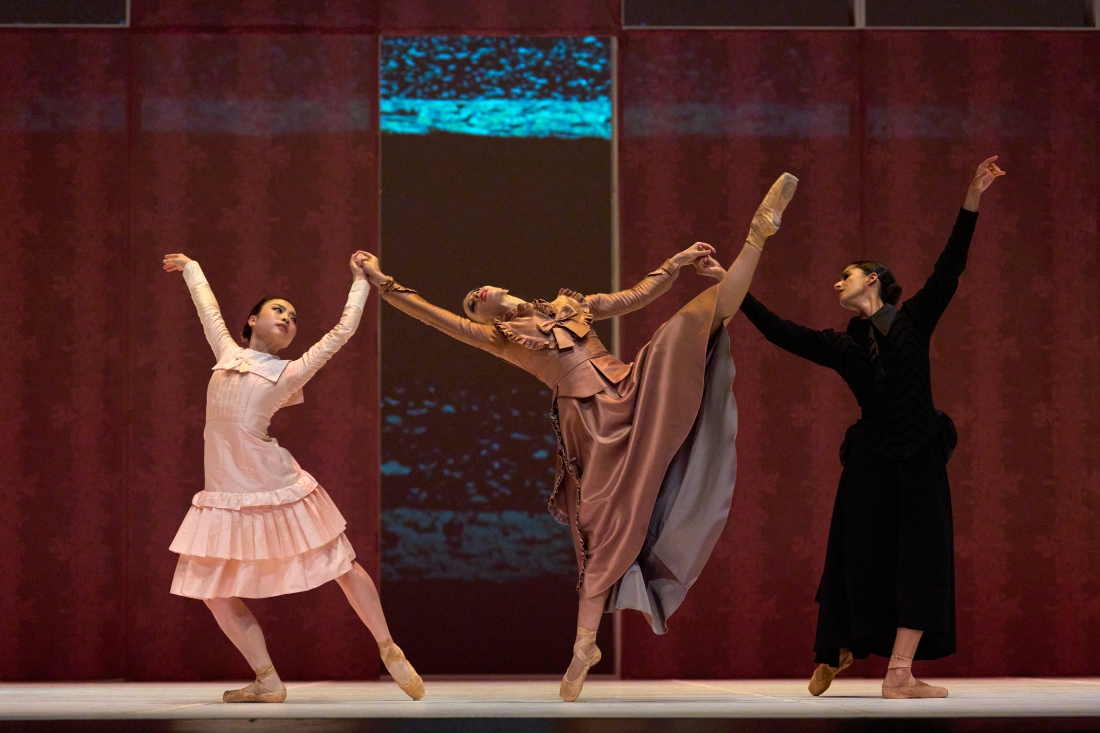 Color stage photograph. Three dancers in contrasting long dresses form a joint composition: the central figure lifts a high leg on pointe while being supported by the other two, who lean in opposite directions.