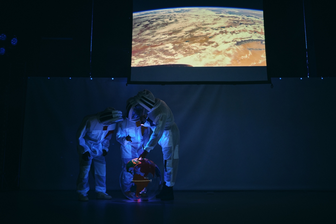 Três pessoas com fatos de proteção iluminam um globo terrestre transparente com pequenas lanternas. Acima delas, uma tela exibe uma imagem aproximada do planeta Terra.