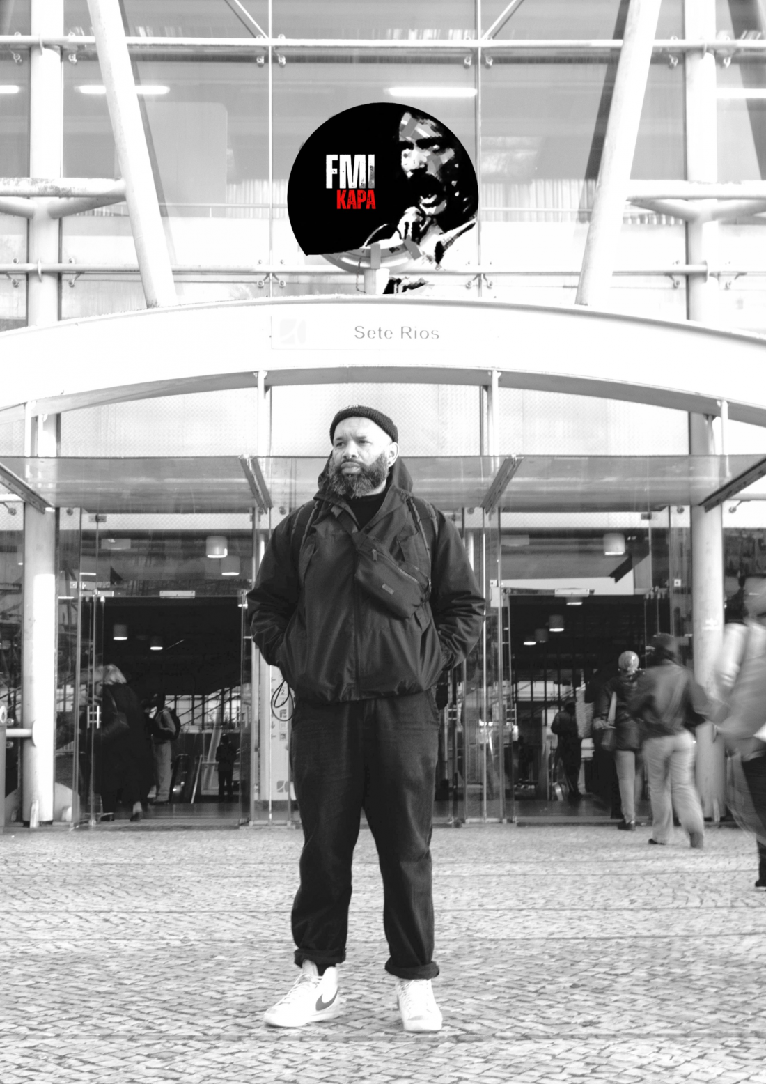 A preto e branco, um homem de pé, com expressão contemplativa, em frente à estação rodoviária de Sete Rios, em Lisboa. Na imagem, vê-se um selo onde se lê “FMI KAPA”.