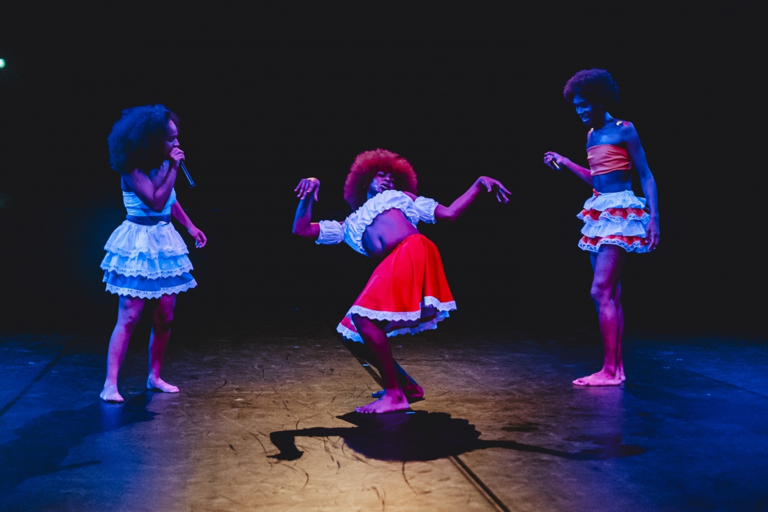 Fotografia de cena a cores. Três intérpretes, iluminadas por luzes em tons de amarelo e azul; ao centro, uma delas dança enquanto as outras duas a observam, cada uma segurando um microfone. Usam saias de folhos e tops em cores vivas.