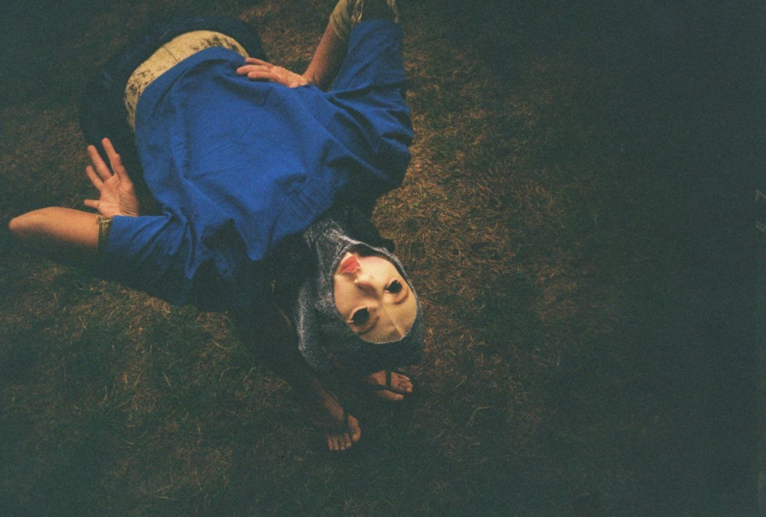 Color photograph in a dark setting. A person dressed in blue and wearing sandals arches their body while wearing a light-colored mask, positioned upside down.