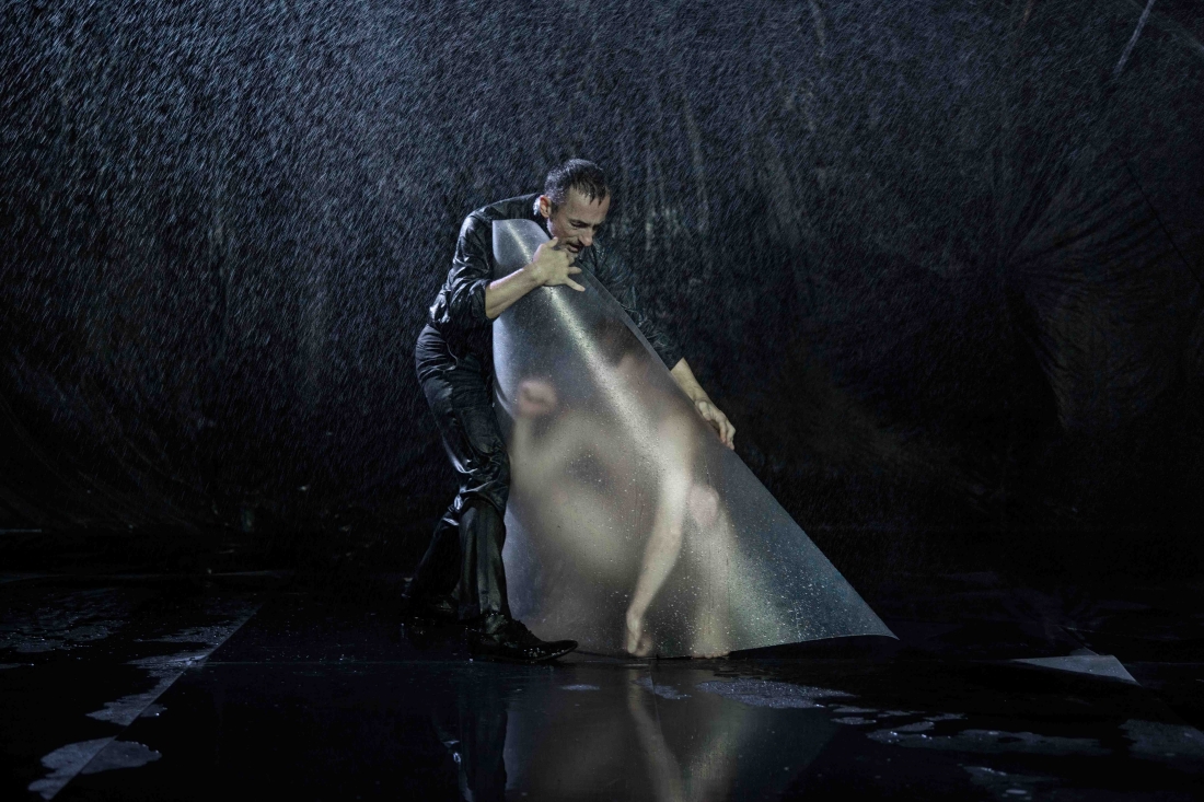 Colour stage photo. In the rain, Dimitris Papaioannou wraps a naked and stooped performer, with a transparent plastic plate.