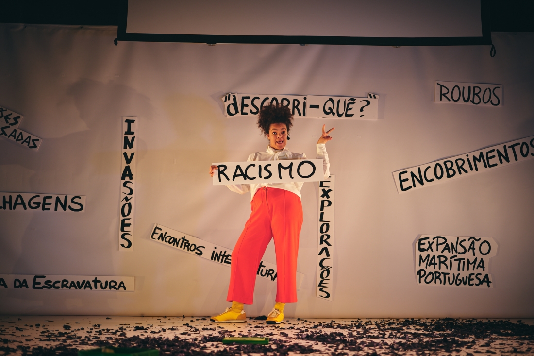 Uma mulher negra fala e gesticula de forma expressiva enquanto segura uma placa de papel branco com a palavra “RACISMO”. Atrás dela, uma tela branca exibe várias placas com palavras como “INVASÕES”, “ROUBOS” e “ENCOBRIMENTOS”.