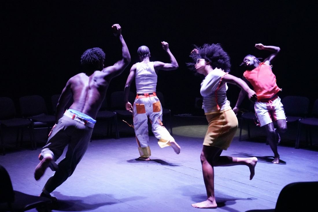 Four performers dance on a circular stage under stage lighting in shades of blue and purple.
