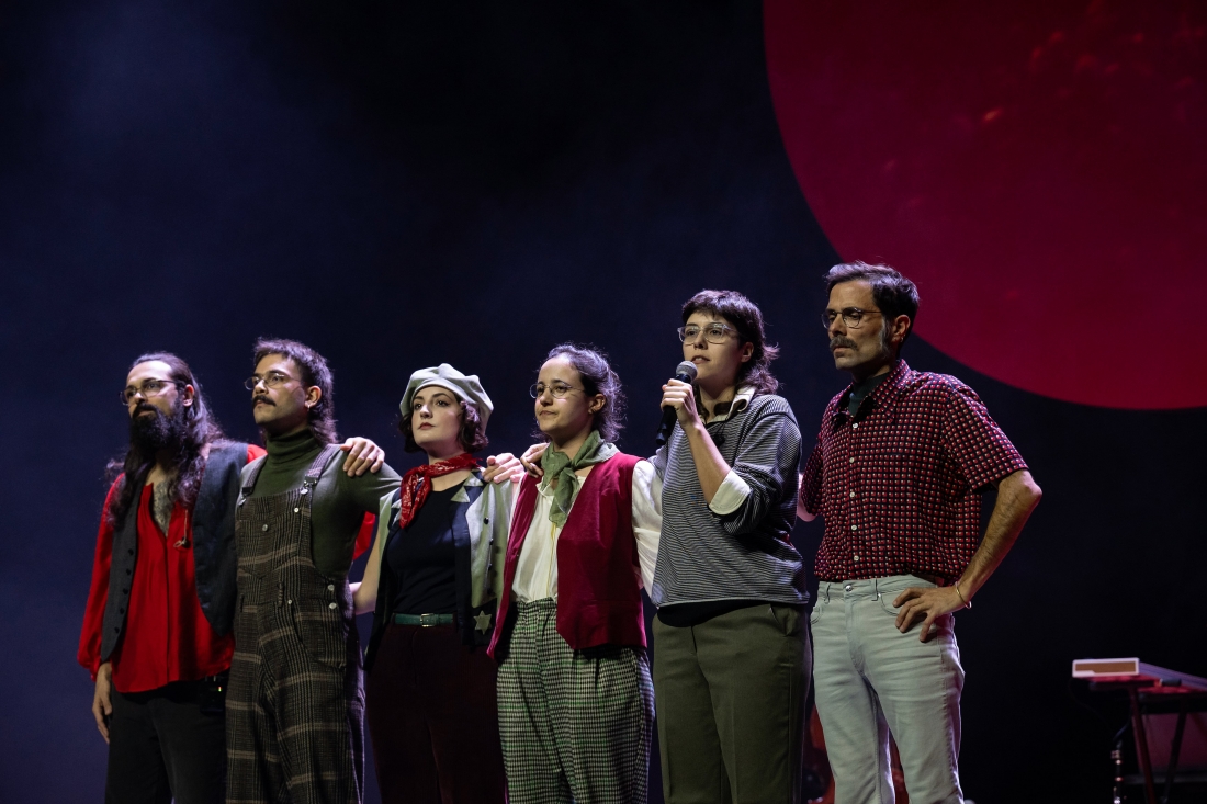 Fotografia a cores de uma pessoa erguendo um cravo vermelho. O cravo destaca-se na fotografia, enquanto a pessoa surge desfocada.