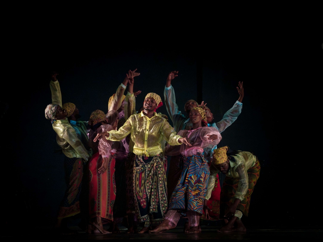 A group of performers on stage, in different positions, in a choreographed arrangement with their arms raised. Their colourful, translucent costumes contrast with the dark backdrop of the scene.