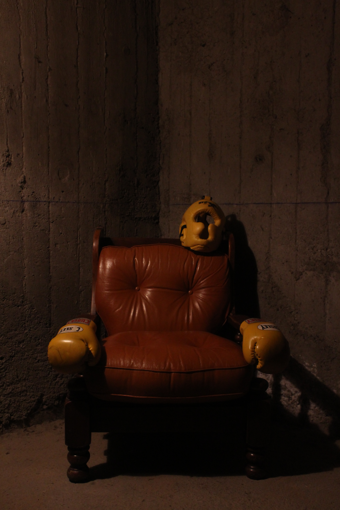 A brown armchair is leaning against a concrete wall, with a helmet and two yellow boxing gloves resting on it.