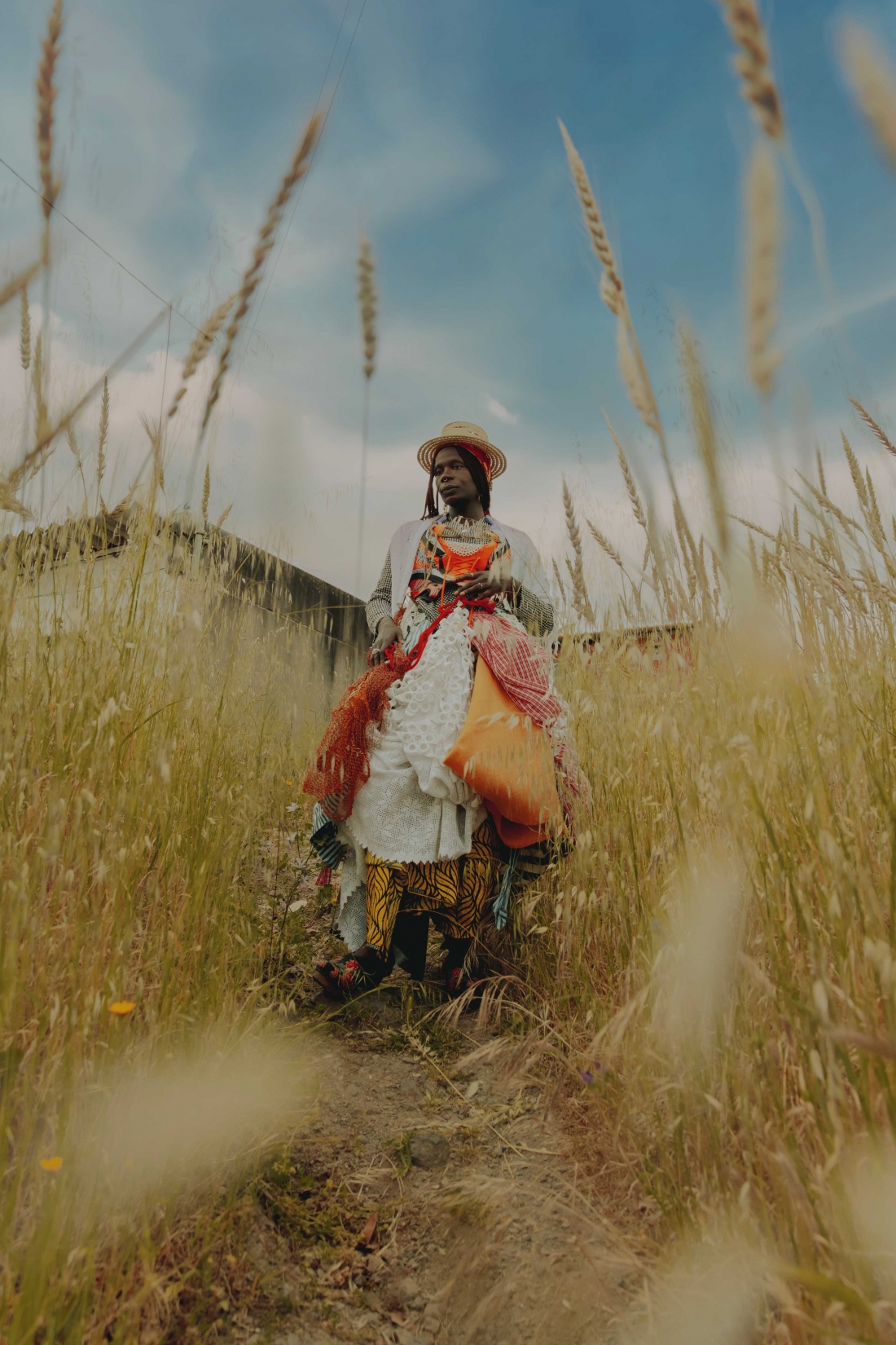 A woman is standing in a wheat field, wearing a straw hat and several layers of clothing in different fabrics and patterns.
