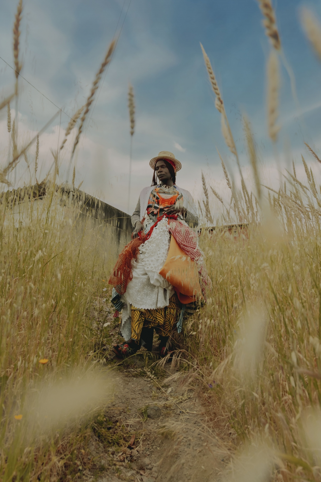 Uma mulher está num campo de trigo, usando um chapéu de palha e várias camadas de roupa com diferentes tecidos e estampados.