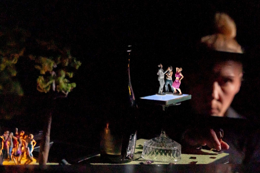 Performer observes miniature figures of people dancing on an illuminated pedestal, whilst other figures appear blurred in the scene.