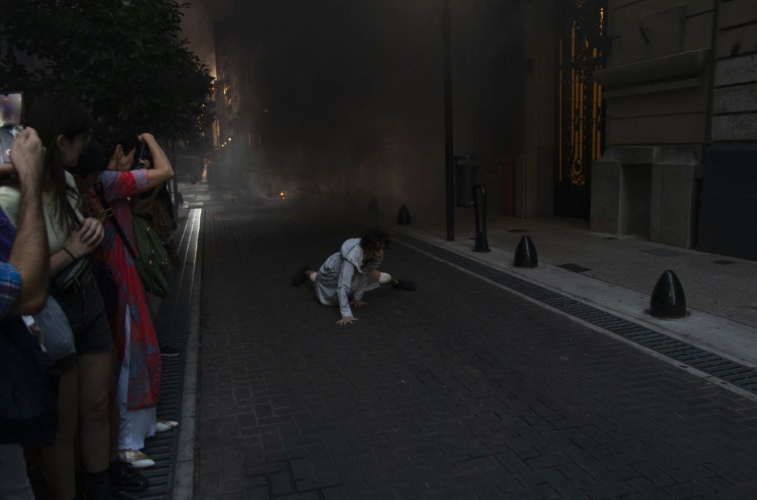 A narrow, dark street; people are taking photos of someone crouching on the ground. Behind them, there is smoke.
