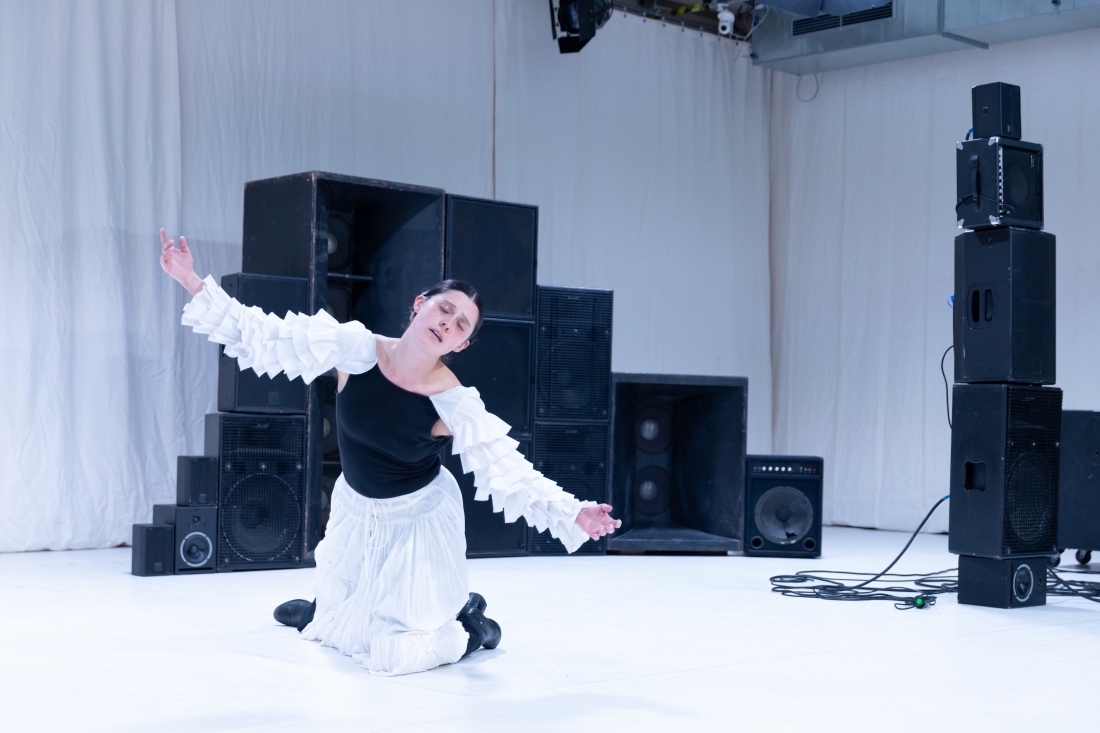 A performer kneeling in a white space stretches out her arms. She is wearing a black and white dress with puffed sleeves, surrounded by several stacked speakers.