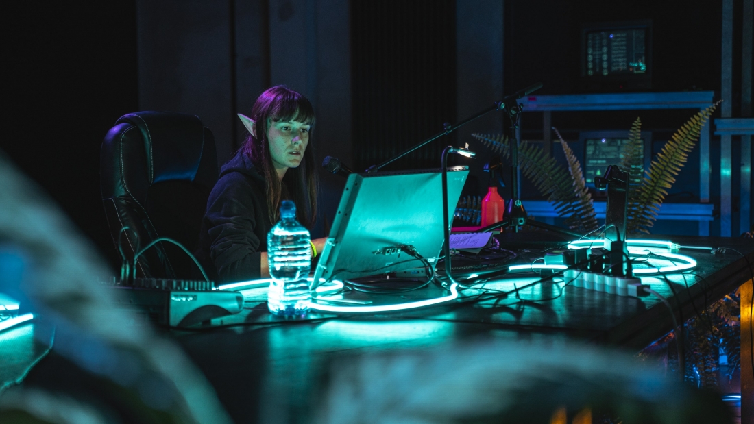 Fotografia de cena a cores mostra Magda Szpecht sentada em frente a um computador. Numa mesa, o computador, vários aparelhos eletrónicos, luzes néon azul e uma garrafa de água.
