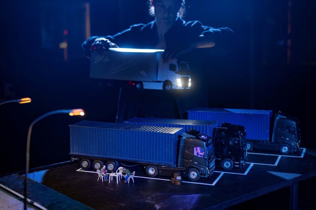 A performer moves a miniature lorry across a model featuring parked lorries.