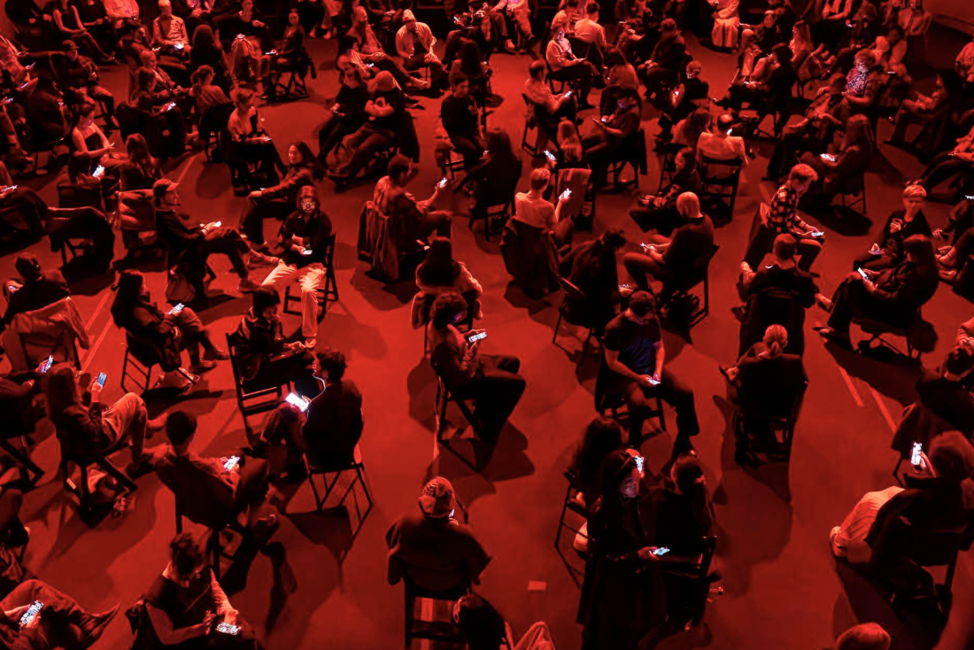 An aerial view of a large group of people sitting on chairs scattered around the space, bathed in red light. Many are holding lit-up mobile phones.