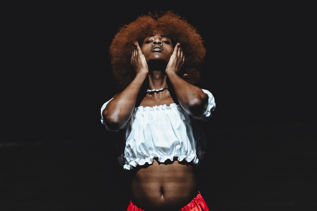 Fotografia de cena a cores. Uma intérprete, iluminada contra um fundo escuro, segura o rosto com ambas as mãos enquanto olha ligeiramente para cima. Usa um top branco e calças vermelhas.