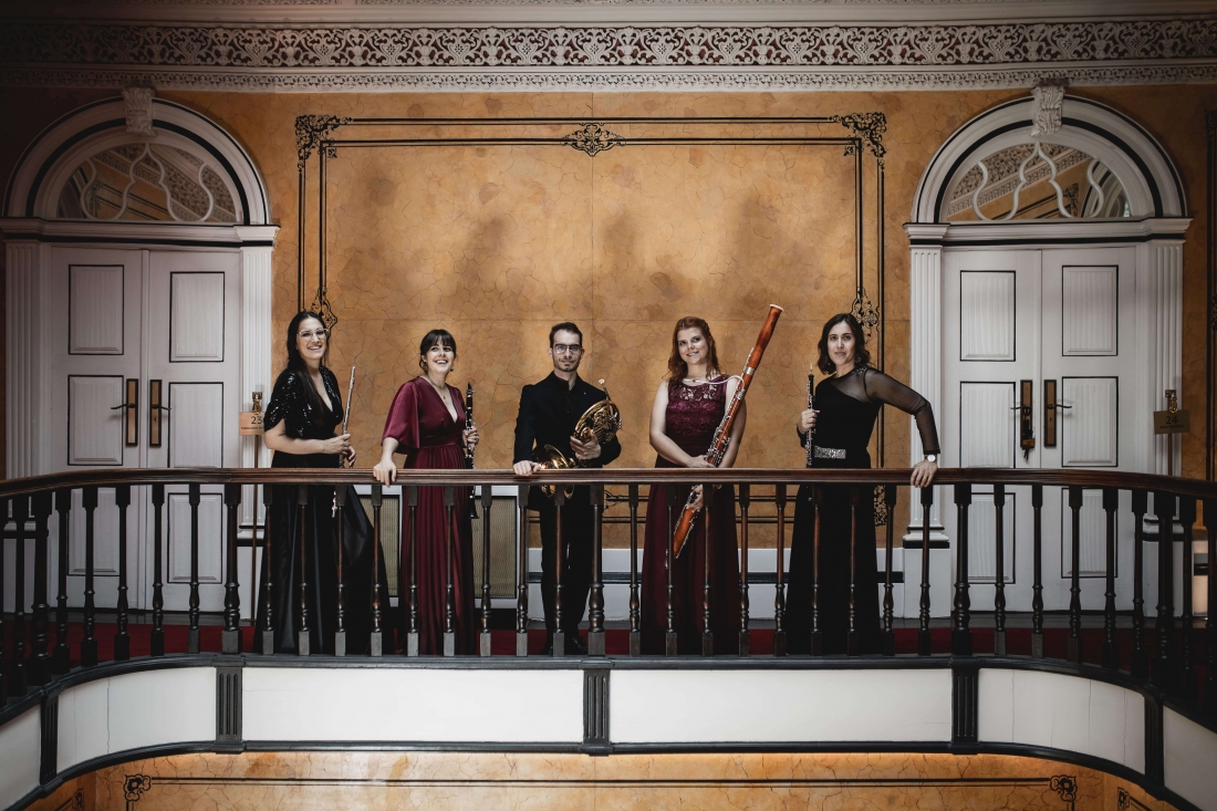 Fotografia a cores mostra Quinteto de Sopros do Vale, com seus instrumentos, no parapeito de um balcão interior. Atrás do grupo, uma parede ornamentada com duas portas brancas e entre elas, uma grande moldura dourada que enquadra o grupo.
