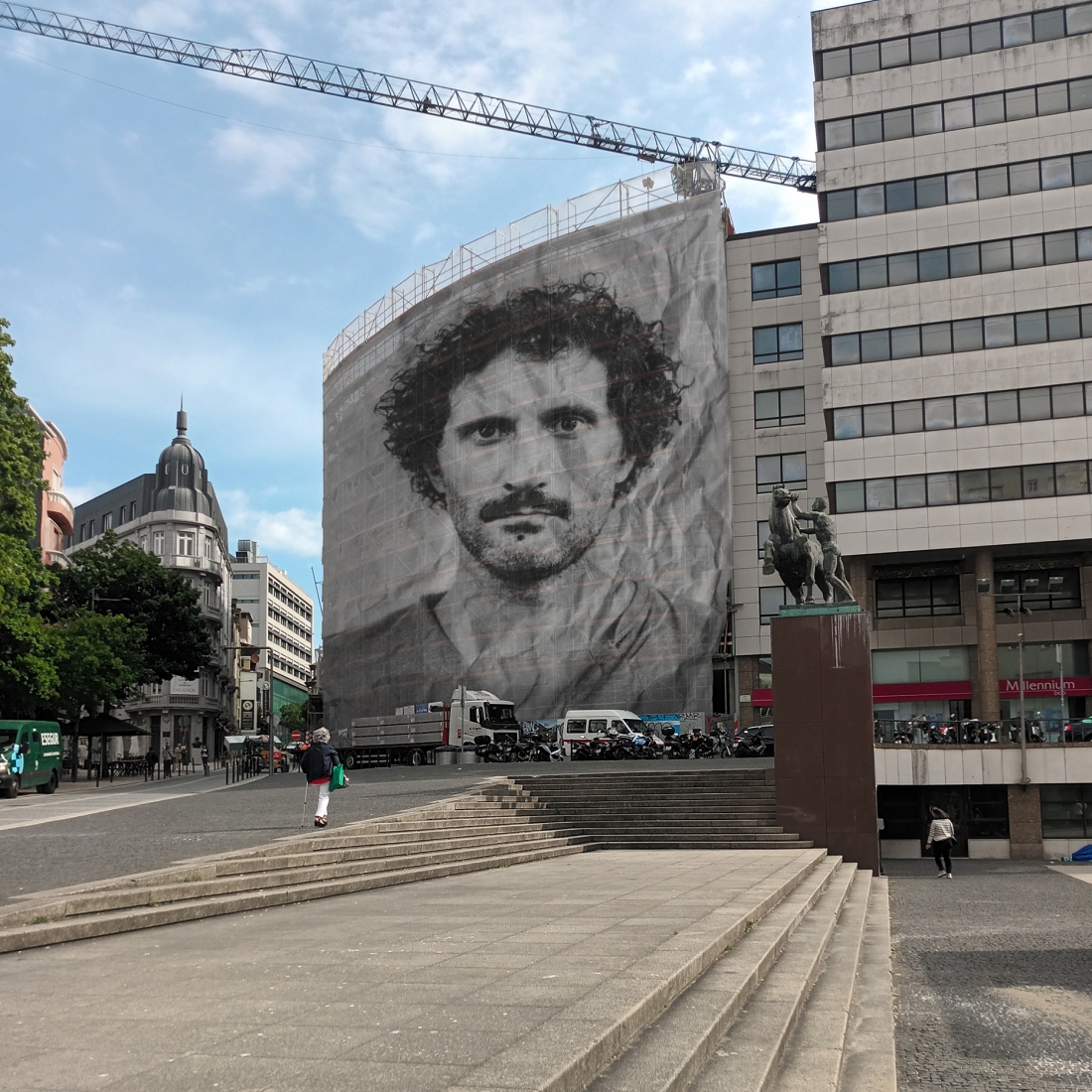 Fotografia a cores mostra a Praça D. João I, em frente ao Rivoli. Um grande retrato a preto e branco de Nuno Preto cobre a fachada de um edifício em construção.