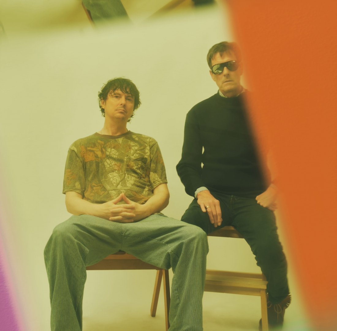 Dois homens estão sentados em bancos de madeira num estúdio de fundo claro, olhando diretamente para a câmara. A imagem é enquadrada por áreas desfocadas em tons de laranja, roxo e verde.