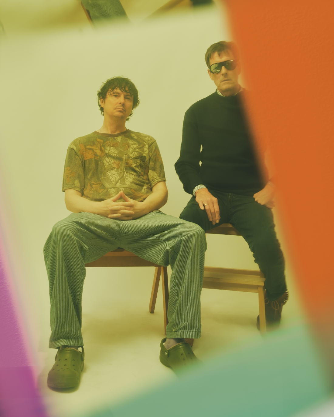 Two men are sitting on wooden benches in a studio with a light-coloured background, looking directly at the camera. The image is framed by out-of-focus areas in shades of orange, purple and green.
