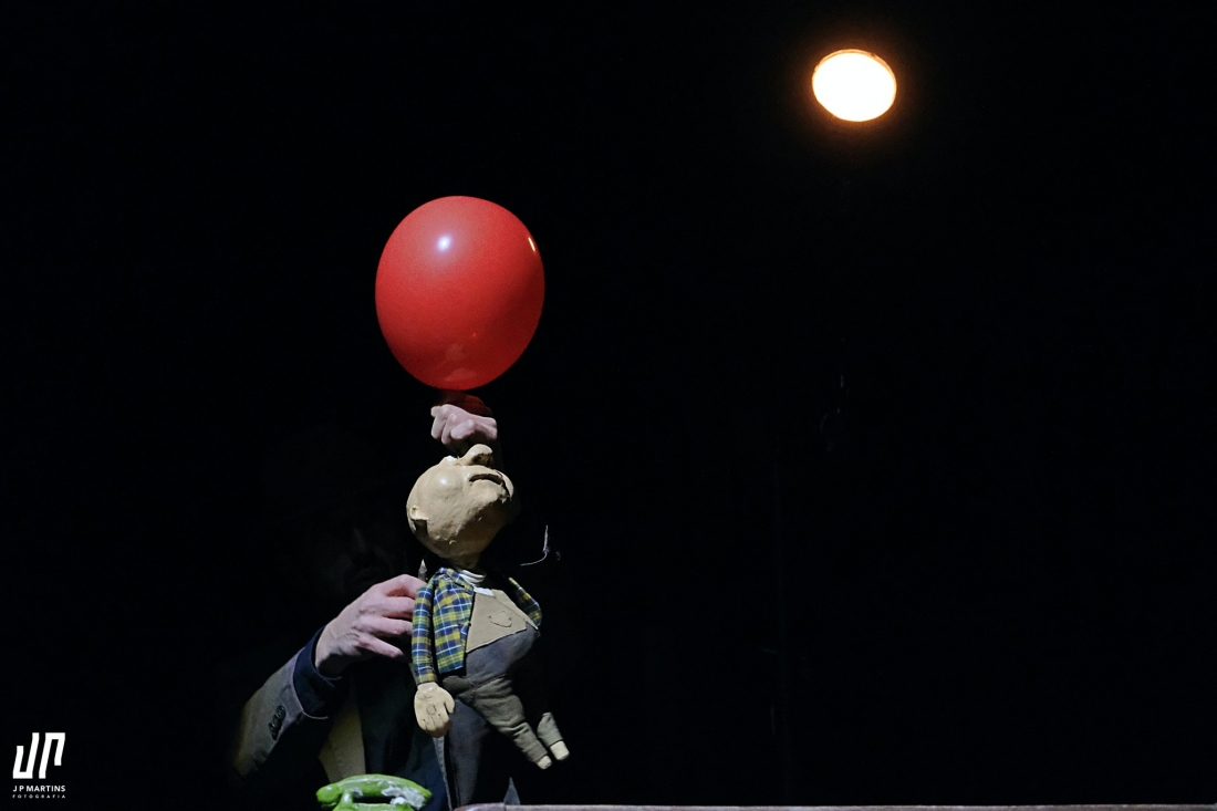 A puppet is held by one hand and is holding a red balloon, illuminated by a spotlight against a black background.