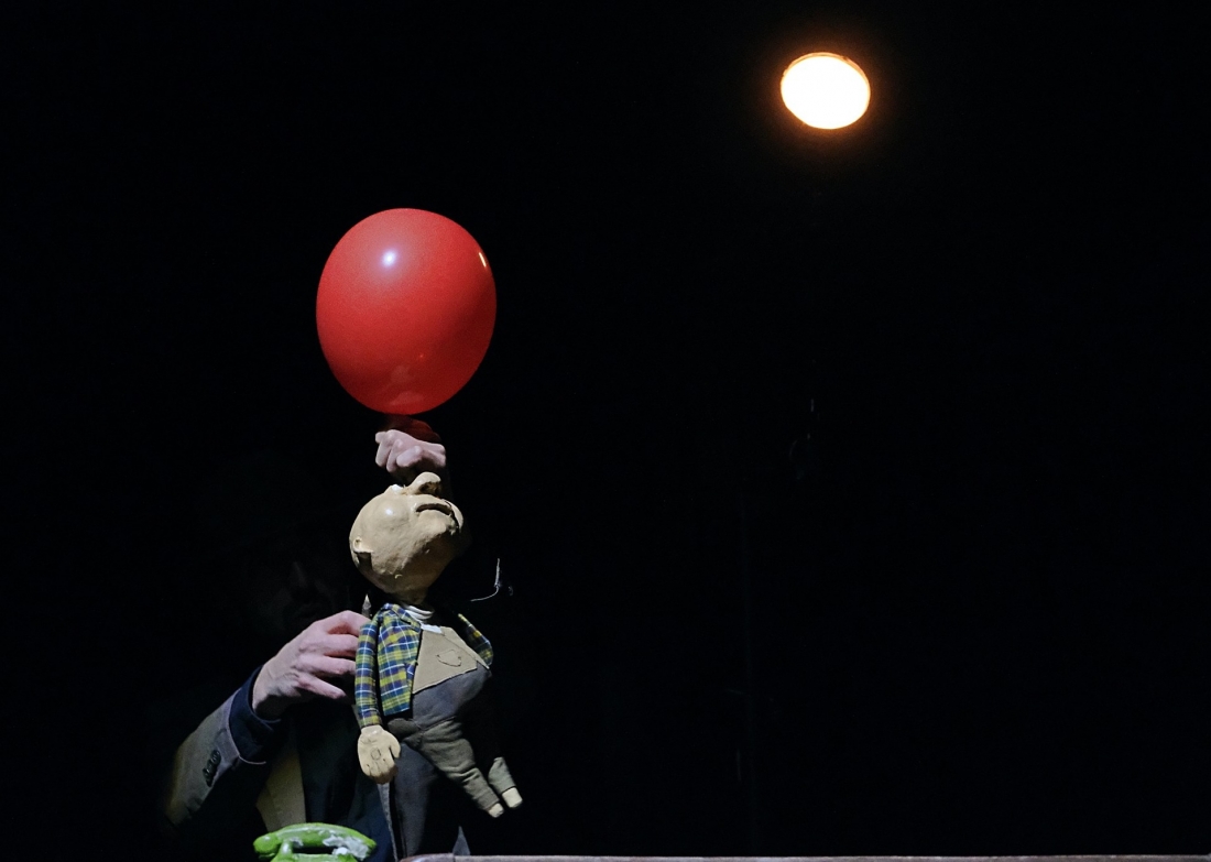 Um boneco é segurado por uma mão e sustenta um balão vermelho, iluminado por um foco de luz sobre um fundo negro.
