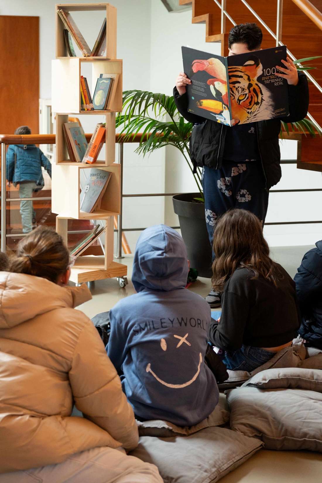 A child is reading a picture book to a group of other children sitting on cushions next to a bookcase. 