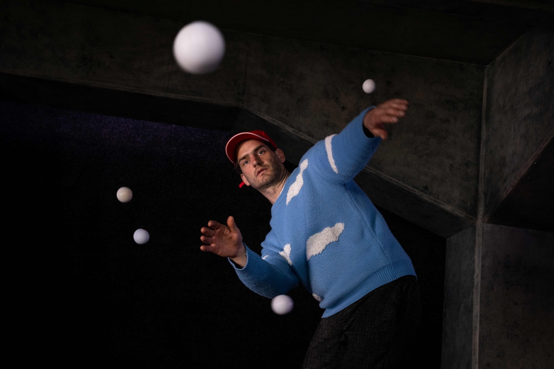 A performer is wearing a blue jumper with white clouds on it and a red cap, whilst juggling several white balls suspended in the air.