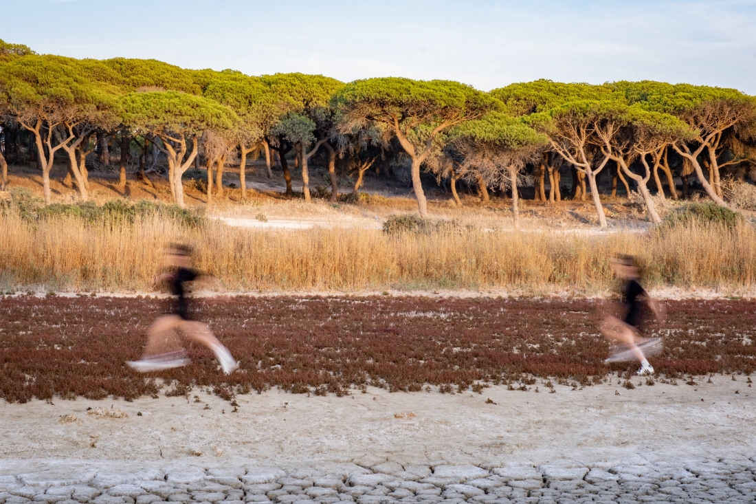 Color photo. In a natural landscape, two people dressed in black and somewhat out of focus run from right to left.