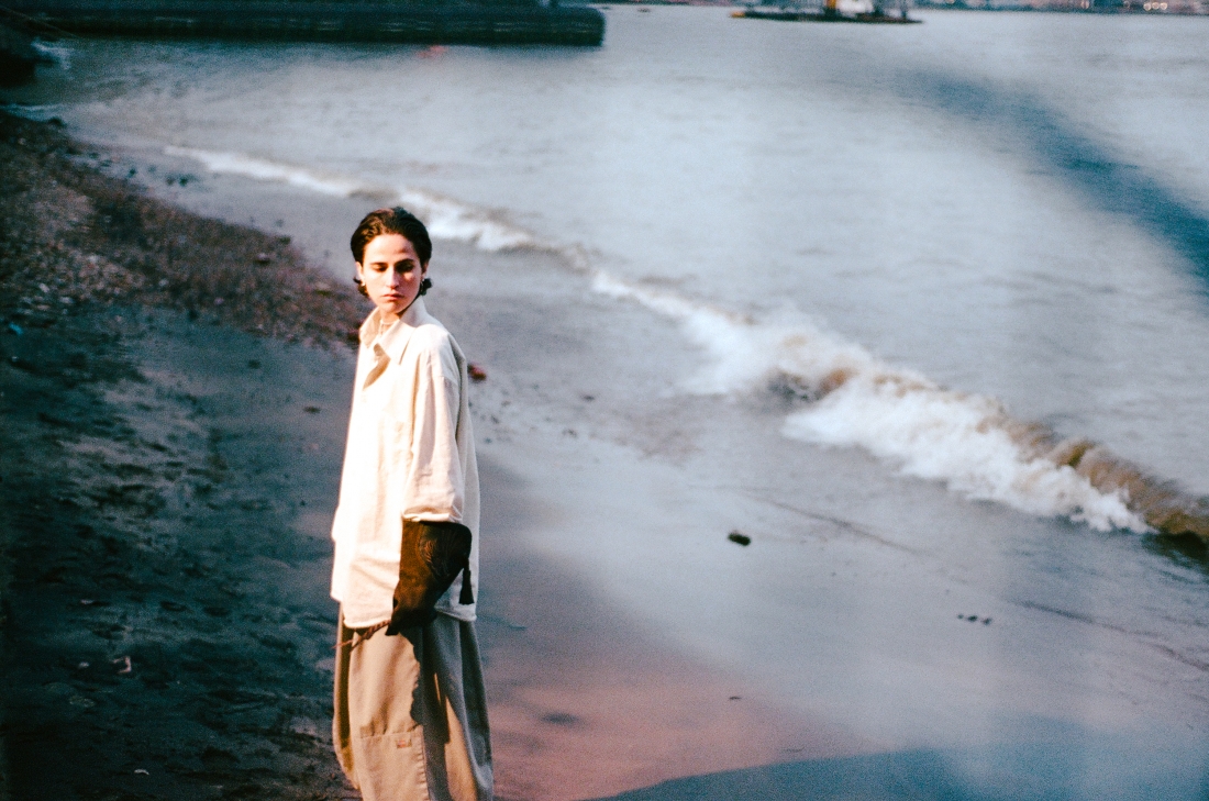 Retrato a cores de Raquel Martins, junto ao mar, de pé, roupas largas, rosto voltado para baixo. 