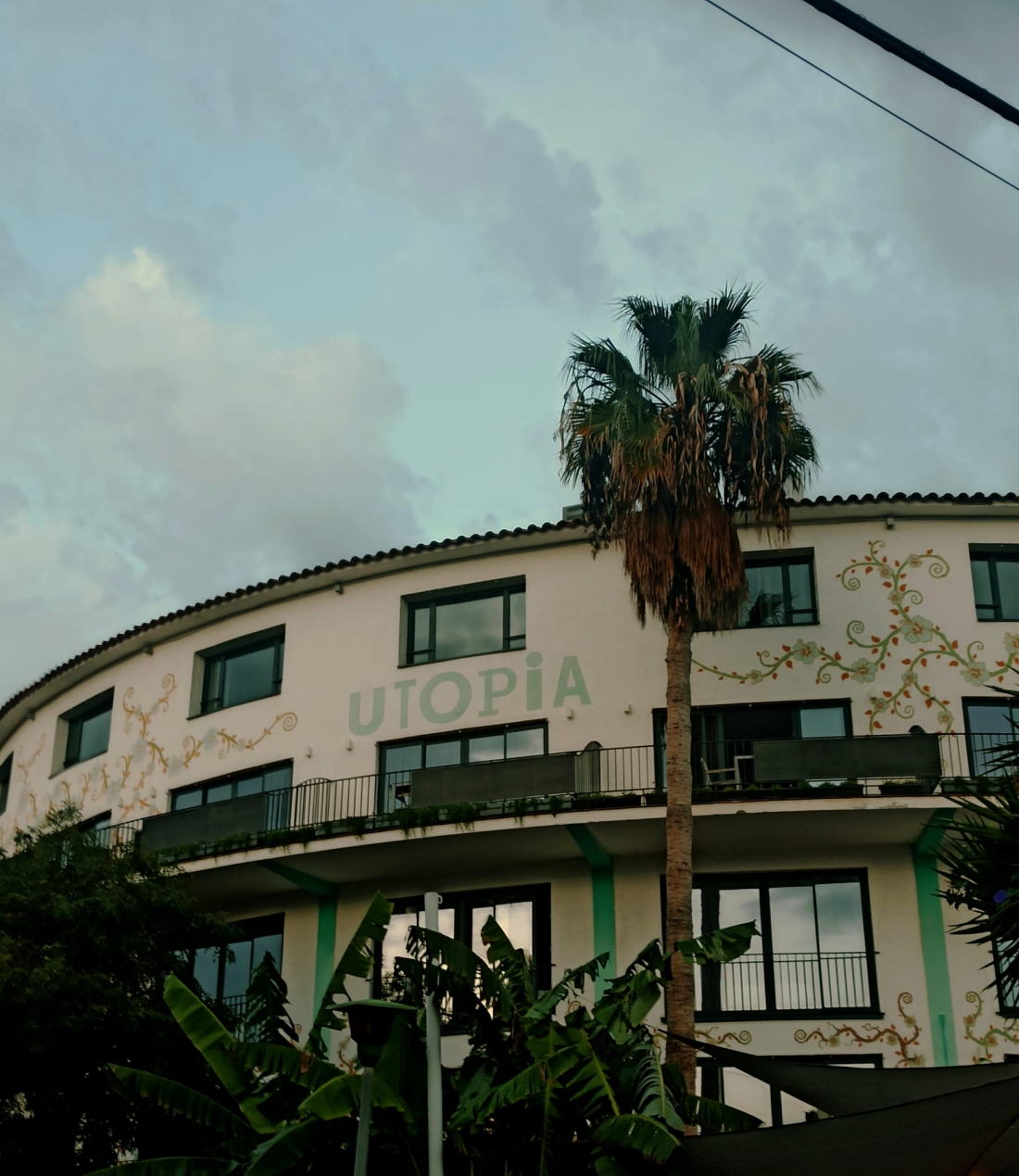 The façade of a building bearing the word ‘UTOPIA’, flanked by a palm tree and tropical vegetation.