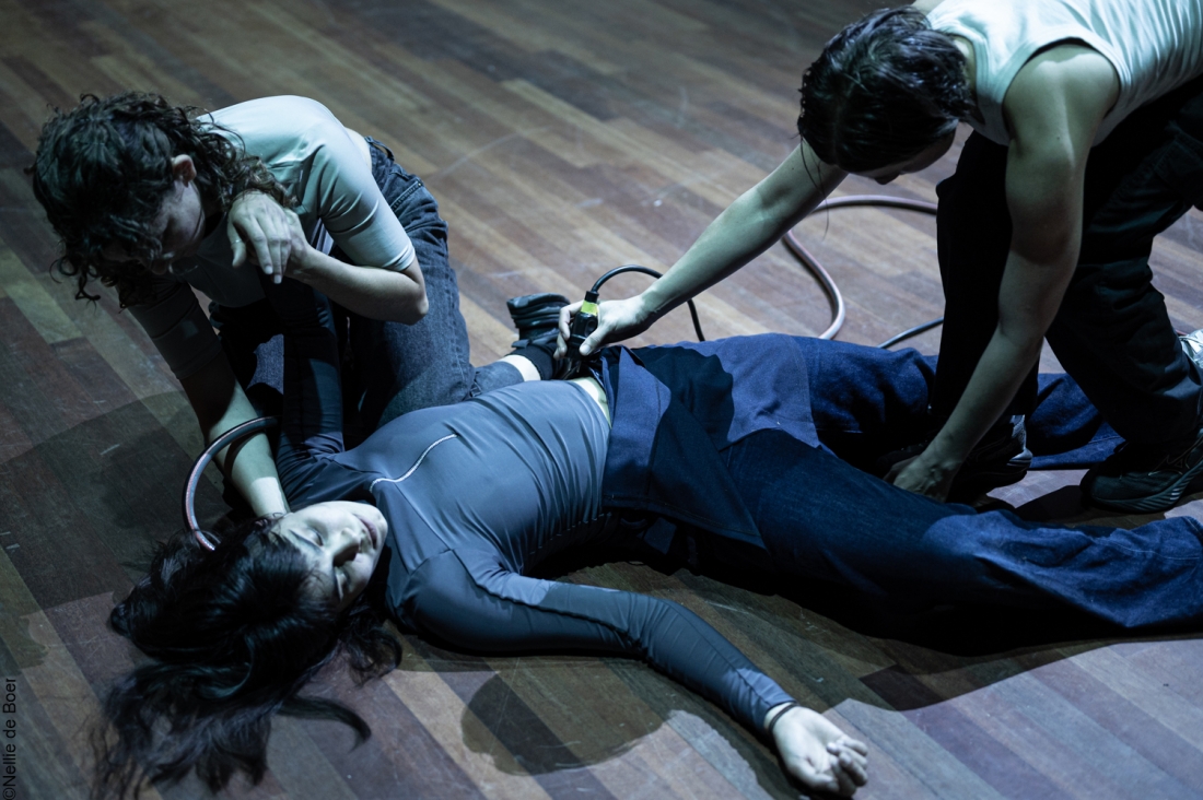 Three people are on a wooden floor, next to a coiled hose, whilst one of them is lying down and the other two are leaning over her.