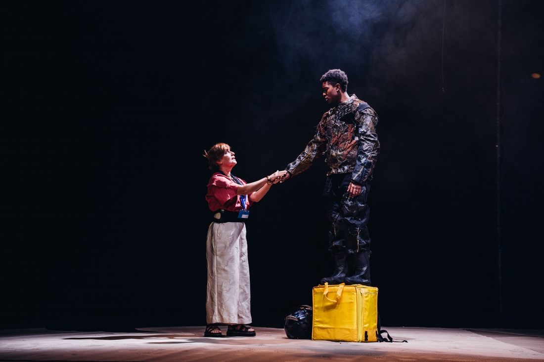 A white woman wearing a blue badge from a European institution is holding hands with a black man, standing on top of a yellow food delivery cooler bag. He and she are looking into each other’s eyes.