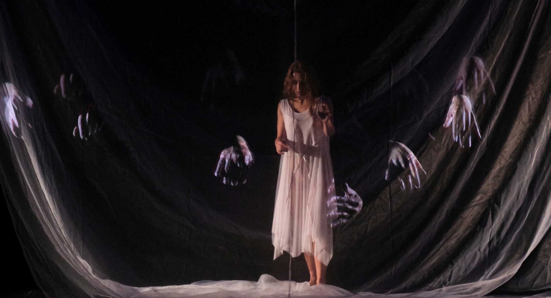 A performer in a white dress stands in the centre of a dark stage, surrounded by translucent fabrics.